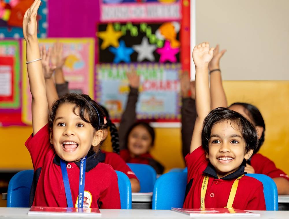 교실에서 의자에 앉아 수업중인 아이들이 한쪽 손을 높이 들고 환하게 웃고 있다