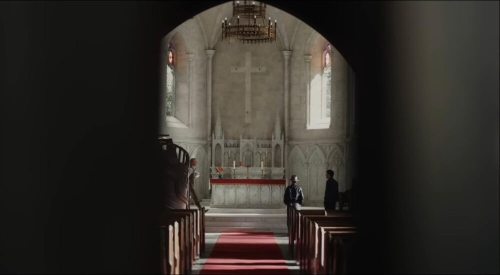 영화 나이브스 아웃3: 웨이크 업 데드 맨 스틸컷 – 제단이 보이는 성당 중앙 통로에 서 있는 세 인물이 포착된 장면