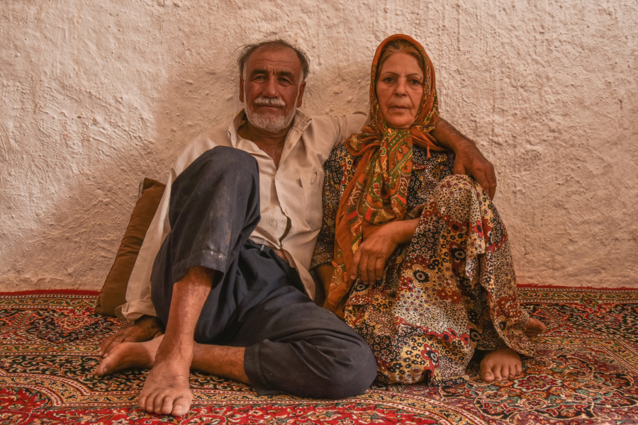 서양의 나이가 지긋한 남자와 여자가 카페트 위에 앉아서 벽에 등을 대고 정면을 응시하고 있는 것을 찍은 사진