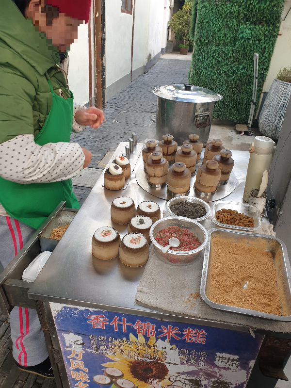 할머니가 쌀떡을 찌고 있다. 나무틀에 쌀떡 재료가 담겨 있고 구매한 2개의 쌀떡에 꼬치를 꼽고 있다.