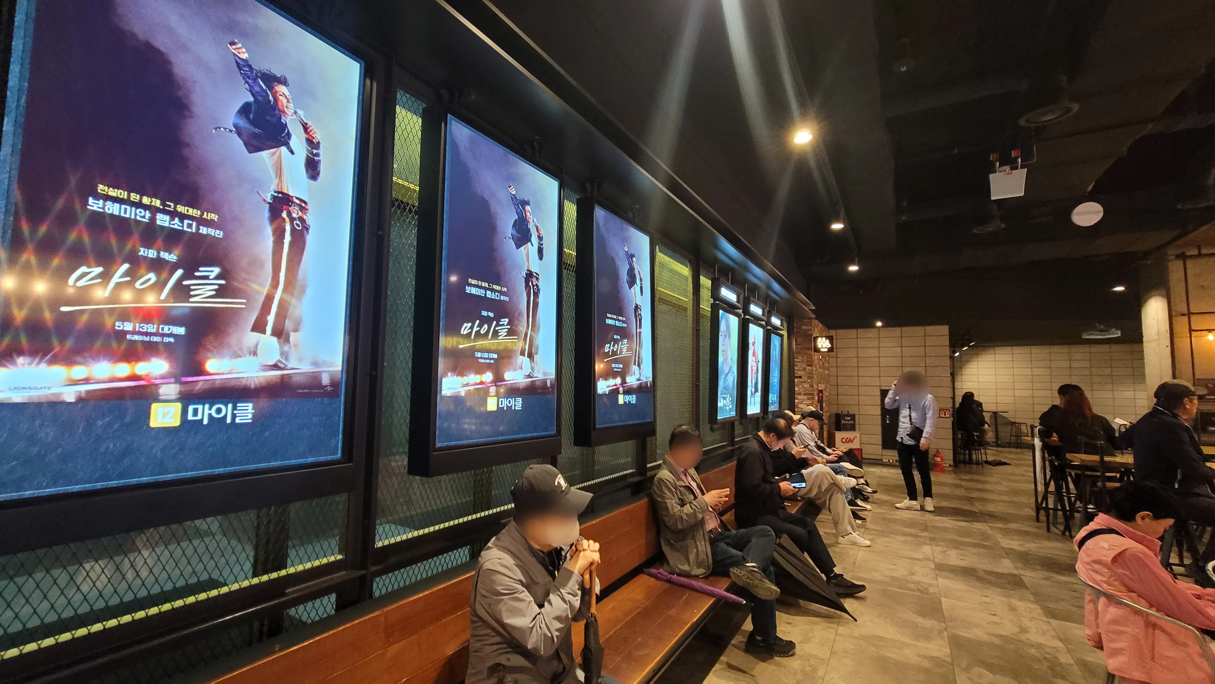 CGV 피카디리1958 극장 내부 대기 공간. 벽면에는 영화 마이클 포스터가 여러 장 걸려 있고&amp;#44; 관객들이 벤치에 앉아 휴대폰을 보거나 대화를 나누며 상영을 기다리고 있다.