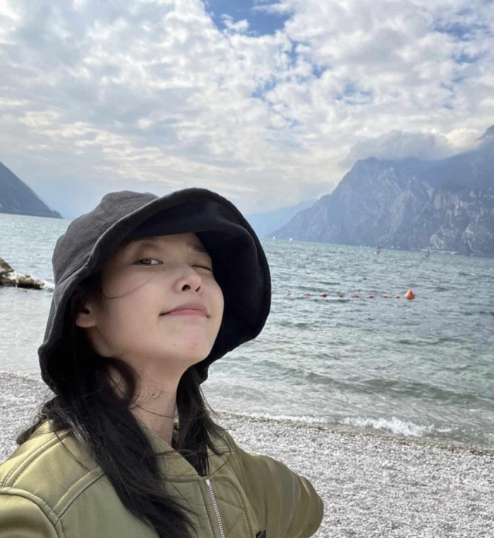 엄청 멋진 호수와 산을 배경으로 챙이 넓은 검은 모자를 쓰고 한쪽 눈을 감은 아이유의 사진