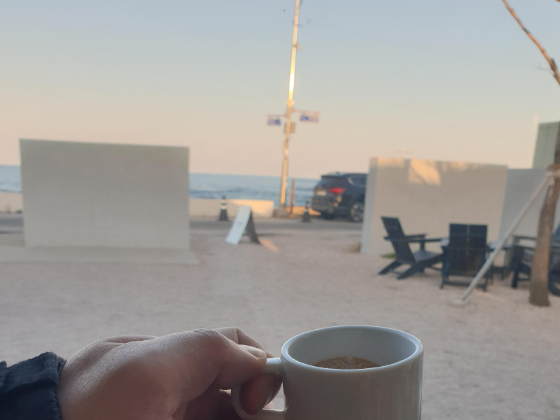 담장 사이로 바다가 보인다. 카페 내부에서 커피 잔을 들고 있다.