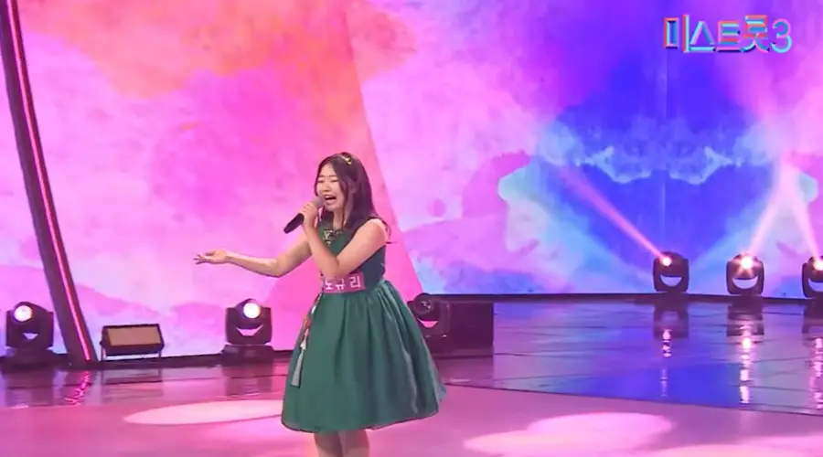 미스트롯3- 초록색 민소매 아래로는 퍼지는 치마를 입고 어깨까지 오는 머리에 금색 머리띠를 하고 왼손으로 마이크를 잡고 오른손은 먼곳을 가리키며 노래하는 노규리
