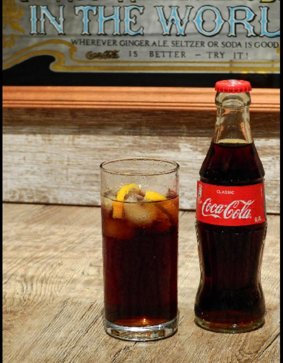 콜라가 물방울이 맺힌 투명 유리컵에 담겨 있고, 그 오른쪽으로는 코카콜라 한 병이 병뚜껑이 닫힌 채로 놓여 있다.