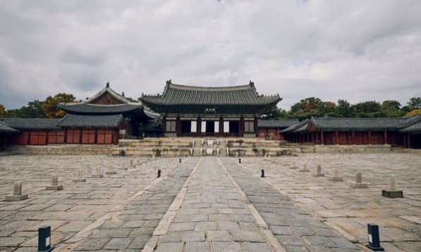 먹구름이 낮게 깔린 하늘 아래 펼쳐진 창경궁 명정전의 웅장한 전경. 전각 앞마당에 줄지어 늘어선 품계석들이 서늘한 분위기를 자아내며, 사도세자의 비극인 '임오화변'이 일어났던 창경궁의 역사적 무게감을 보여주는 사진.