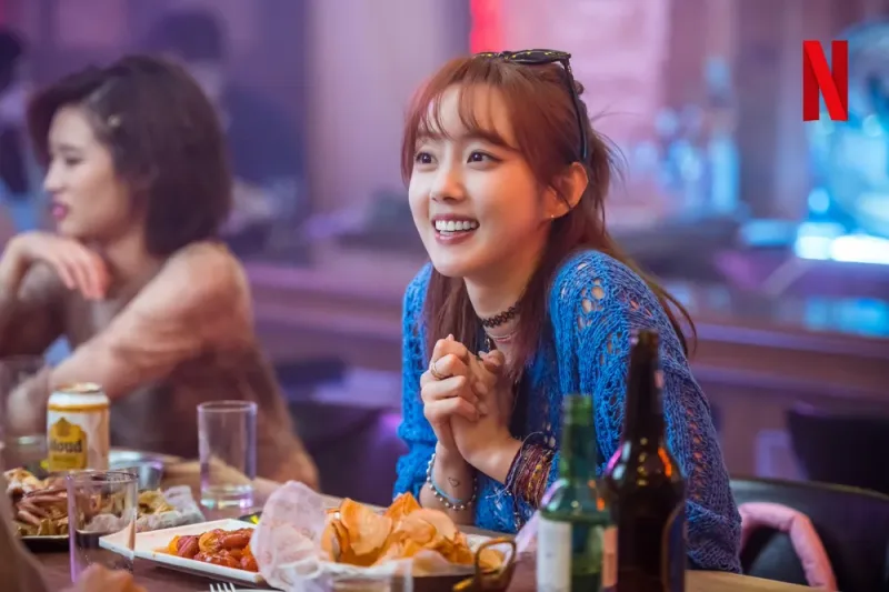 어느 술집을 배경으로 갈색 긴머리에 파란 티를 입고 앉아 있는 드라마 이두나에서 최이라 캐릭터