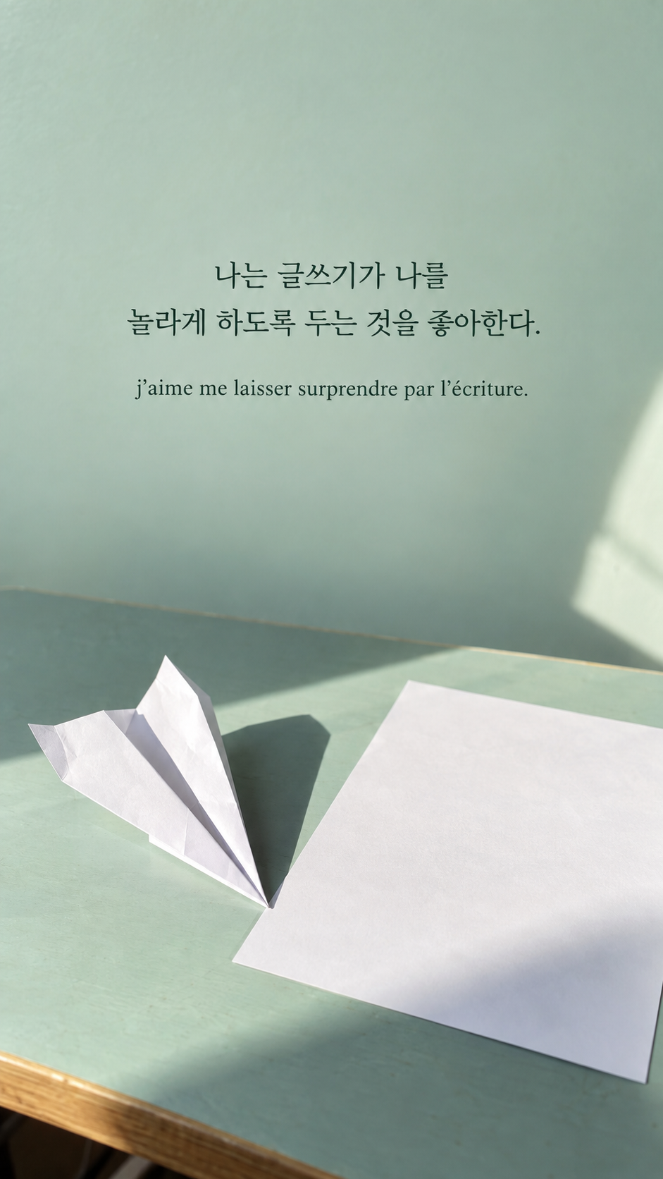 민트색 책상 위 종이비행기 배경에 담은 기욤 뮈소의 말: 나는 글쓰기가 나를 놀라게 하도록 두는 것을 좋아한다.