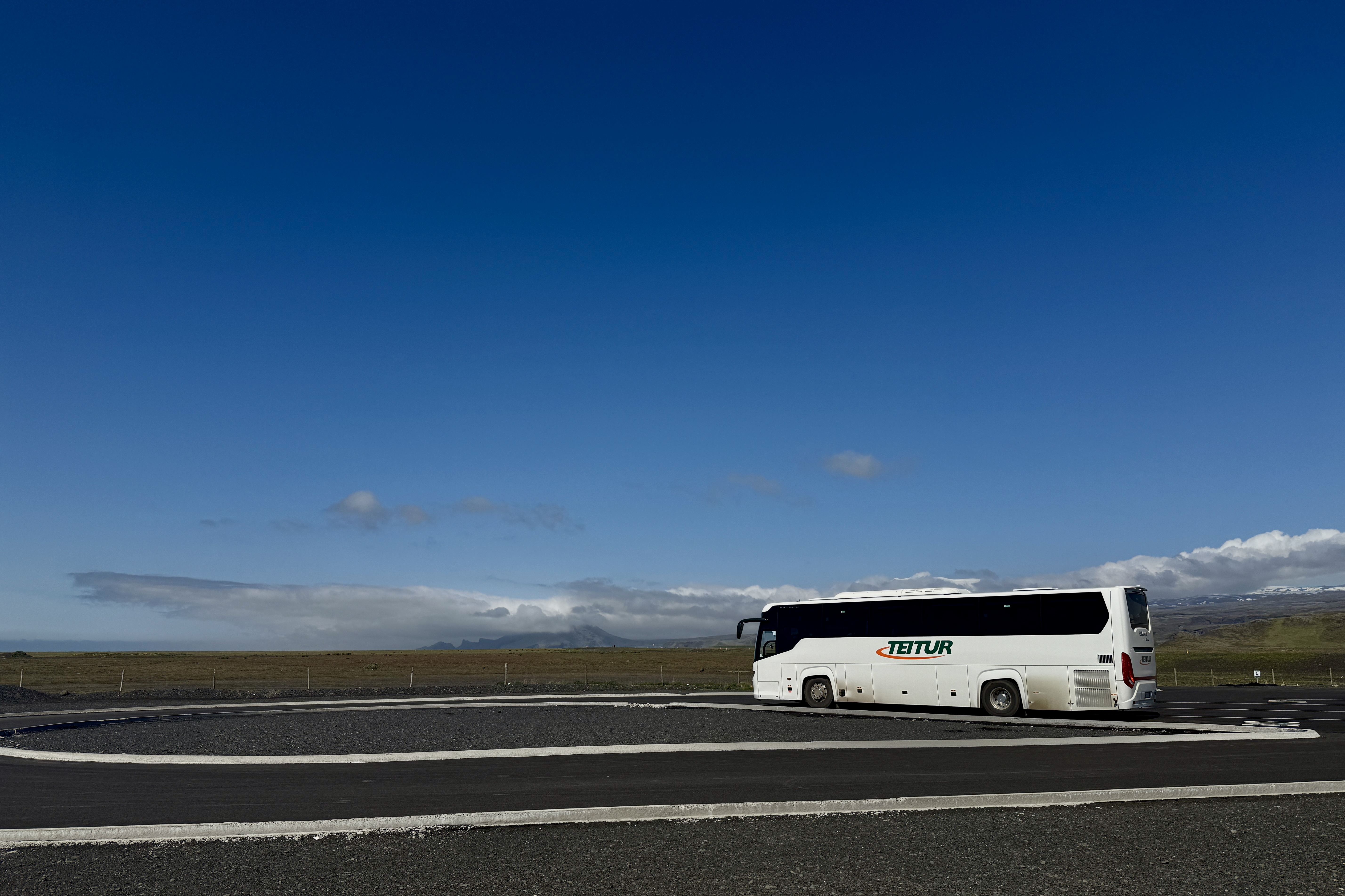 넓고 황량한 아이슬란드 풍경에 하얀색 'TEITUR' 관광버스가 주차장에 서 있는 모습입니다. 배경에는 푸른 하늘과 멀리 구름과 산맥이 보이며, 검은 화산재 지형이 펼쳐져 있습니다.
