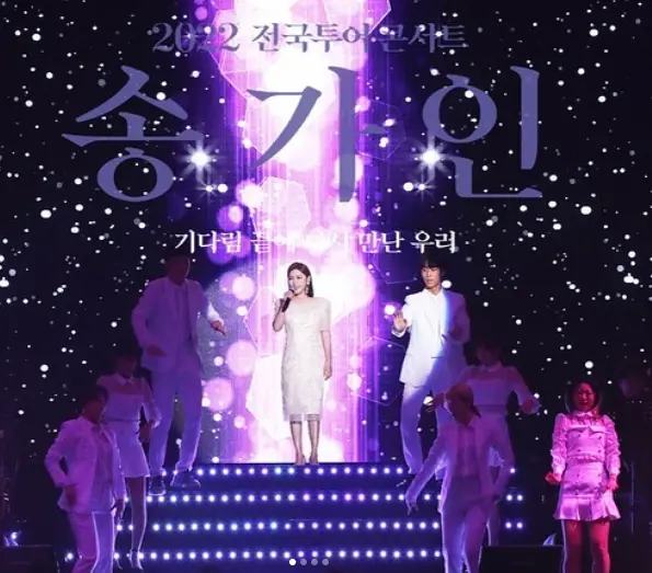 송가인- 검은 무대 불이켜진 계단 위 흰옷을 입고 오른손으로 마이크를 든채 노래하는 송가인과 그옆 흰정장을 입은 댄서들