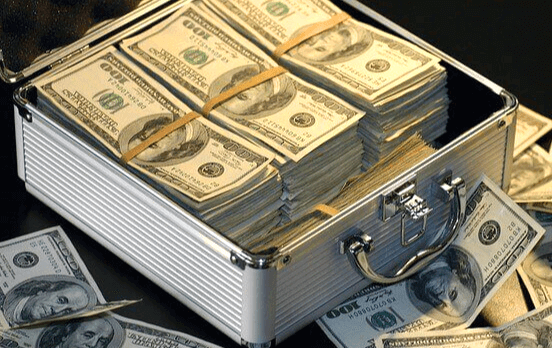은색 돈가방에 달러 지폐가 한 가득 담겨있고, 가방 밖에는 달러 지폐들이 흩어져 있다.