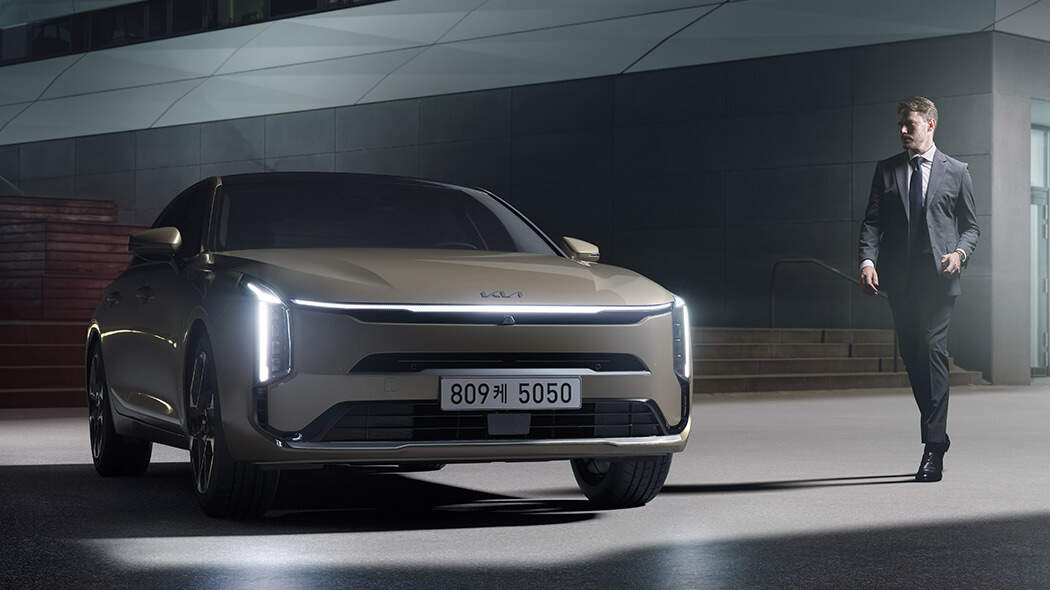 헤드라이트가 켜진 기아 K8 전면, 차량 좌측에 서있는 수트를 입은 성인 남성 1명