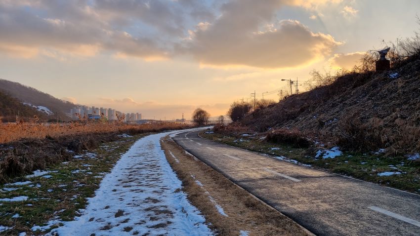 눈 녹은 자전거 길, 왼쪽 산책길에 쌓인 눈, 구름 낀 하늘에 물들기 시작하는 해 질 무렵의 노을, 왼쪽 멀리 보이는 아파트 단지, 그 뒤로 야산,