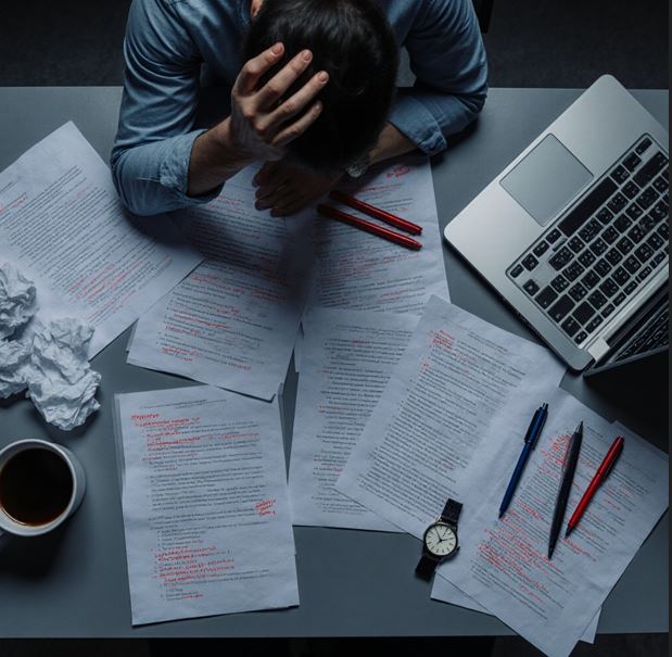 책상 위에 여러 개의 수정된 문서, 구겨진 종이, 붉은색 펜 표시가 가득 하고, 머리를 감싸 쥐고 있는 사람의 모습