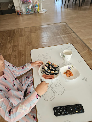 잠옷을 입은 아이가 거실에 앉아 김밥과 김치를 맛있게 먹고 있는 모습.