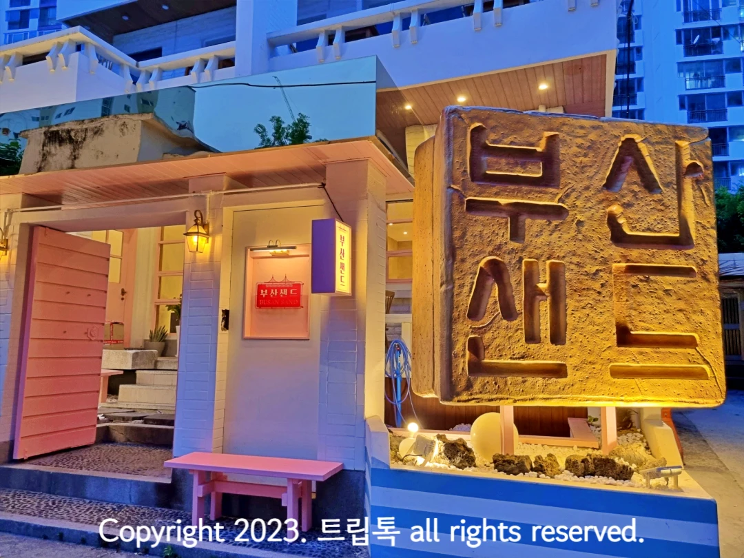왼쪽 나무 대문이 열려있고 오른쪽에 커다란 네모에 부산샌드라고 적혀있다.