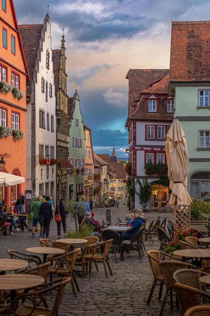 독일 로텐부르크의 구시가지 거리 풍경으로, 형형색색의 목조 건물들이 줄지어 있고, 거리 곳곳에는 카페 테이블과 사람들이 여유롭게 앉아 있는 모습이 담긴 사진. cobblestone 길과 붉은 지붕이 중세 감성을 그대로 전달합니다.