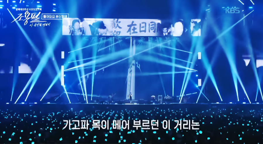 ‘가고파 목이 메어 부르던 이 거리는’ 자막과 함께 파란 조명이 비추는 무대. 조용필이 긴 마이크 스탠드를 잡고 노래하는 장면.