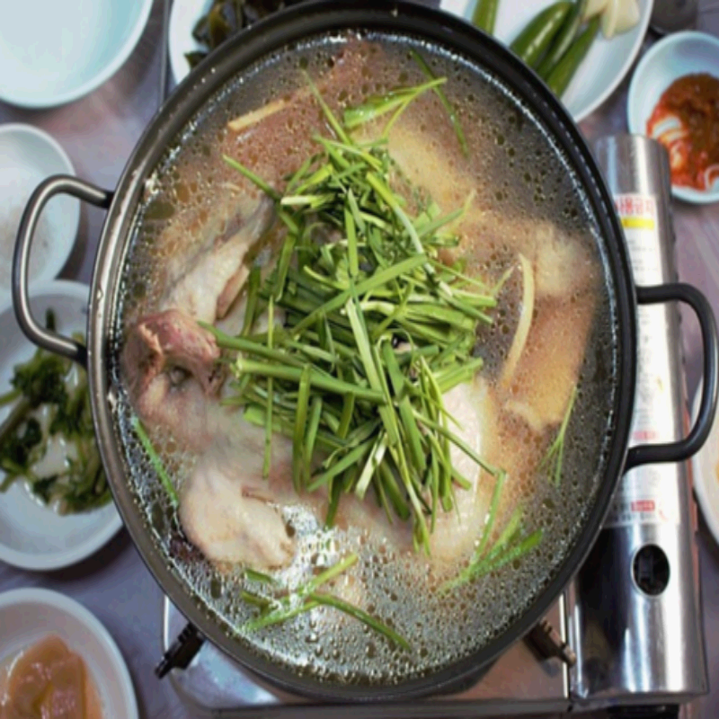 한국 전통 보양식인 삼계탕이 보글보글 끓고 있는 모습입니다. 뚝배기 냄비에 통닭이 그대로 들어가 있으며, 위에는 파와 부추 같은 신선한 채소가 듬뿍 올려져 있습니다. 맑은 국물은 인삼, 대추, 마늘 등이 우러나 건강한 맛을 더해주며, 주변에는 김치, 장아찌, 고추, 양념장 등 다양한 밑반찬이 함께 차려져 있어 푸짐한 한 상 차림을 보여줍니다.