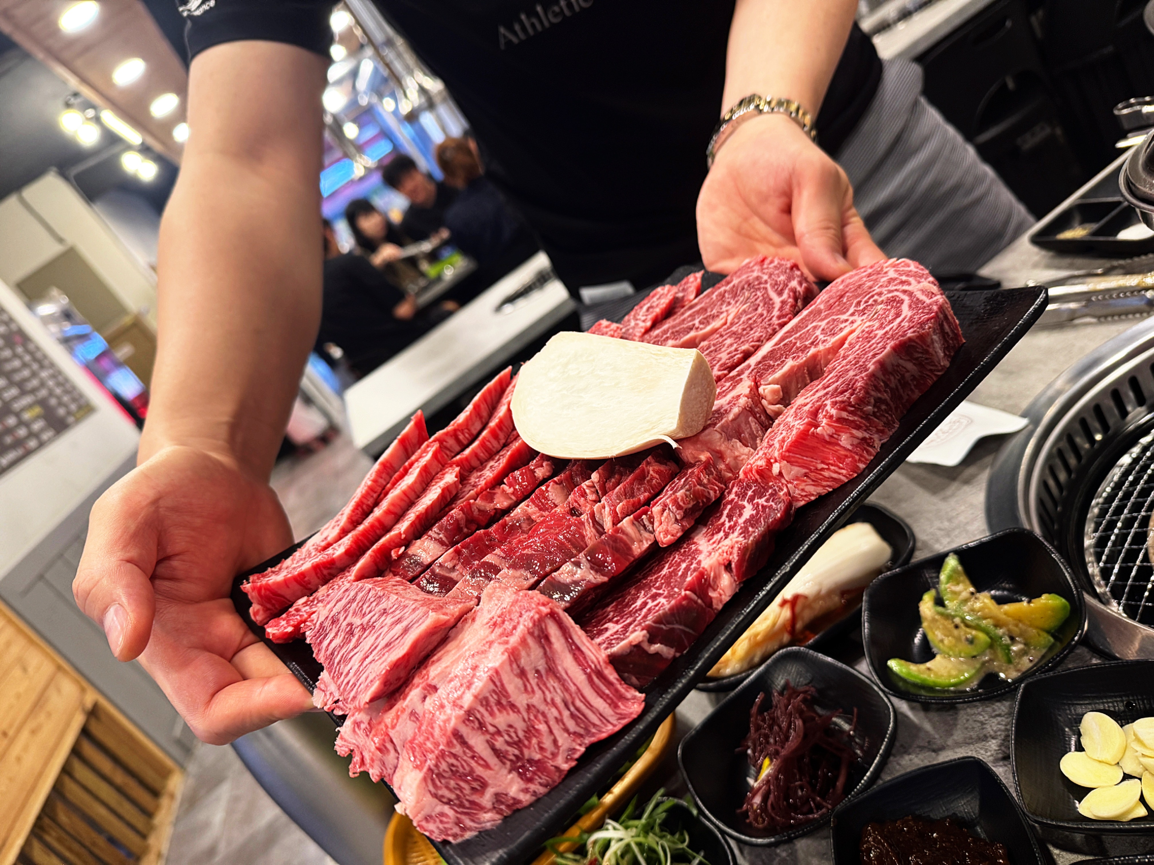 삼거리 명품한우에서 친구가 손으로 들고 보여주는 고기 모둠. 다양한 부위의 마블링 고기가 담겨 있으며 주변에는 반찬 세팅도 되어 있다.