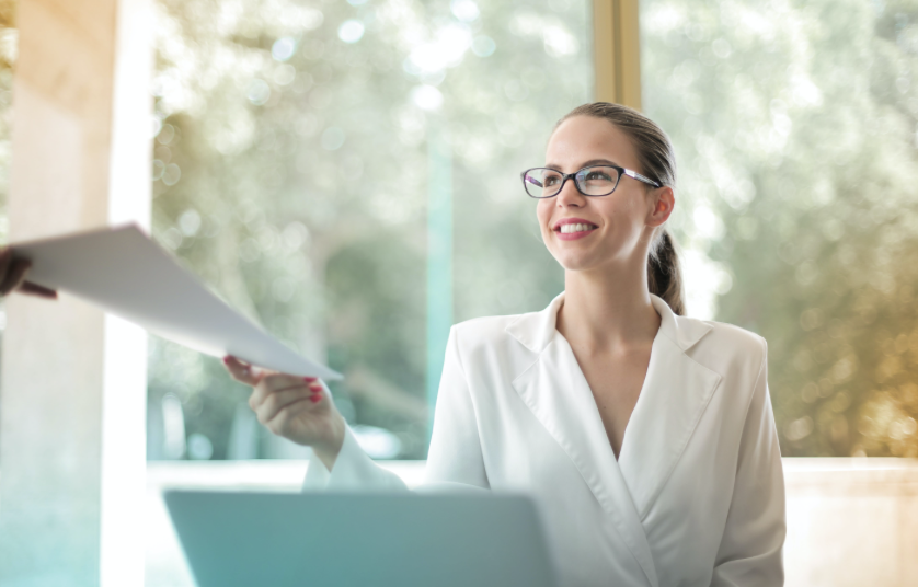 흰 재킷을 입고 검은 뿔테 안경을 쓰고 미소지으며 서류를 넘겨주고 있는 여성