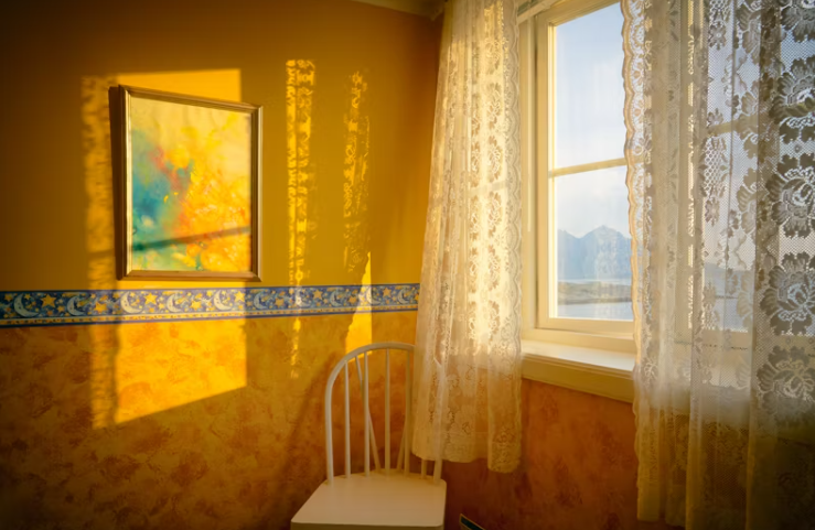 따뜻한 아침 햇살이 벽을 물들이는 조용한 방 안, 흰 의자와 커튼 너머로 들어오는 창가 풍경