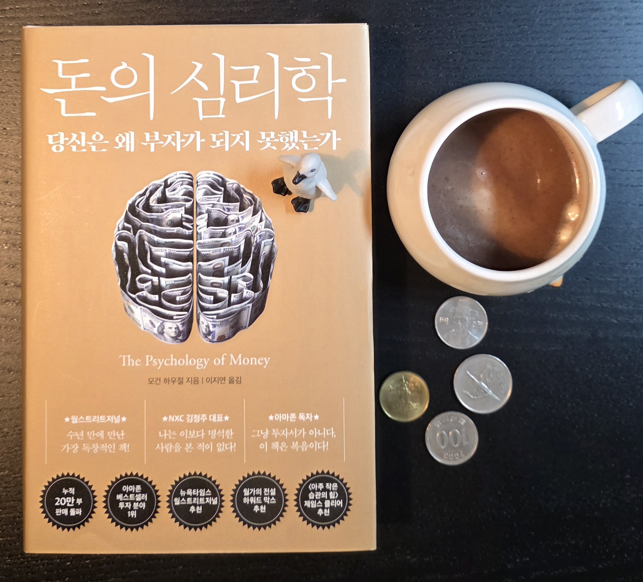 커피 한 잔과 동전, 펭귄 피규어가 함께 놓인 돈의 심리학 책 표지