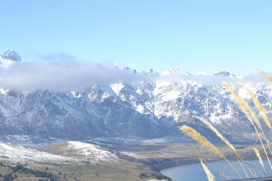 뉴질랜드 남섬 퀸스타운의 와카티푸 호수와 만년설이 덮인 리마커블스(The Remarkables) 산맥 전경. 푸른 하늘 아래 구름이 걸린 설산과 평화로운 호수 풍경.