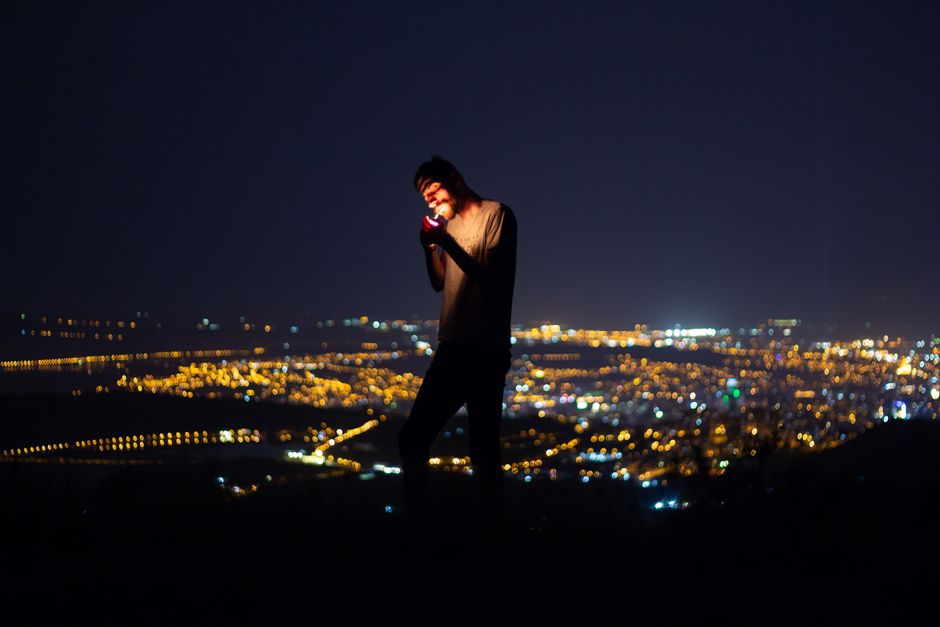 A man lights a cigarette against a vibrant nighttime cityscape backdrop, capturing urban nightlife.