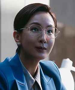 어느 실내에 숏컷 머리와 금테 안경을 쓰고 파란 정장을 입고 앉아 있는 드라마 유괴의 날 모은선 캐릭터