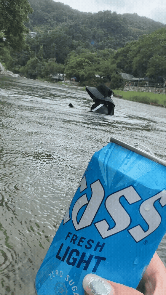 흐르는 계곡물 위로 카스 라이트 맥주 캔을 든 장면. 손톱 네일과 함께 시원한 여름 감성이 돋보인다.