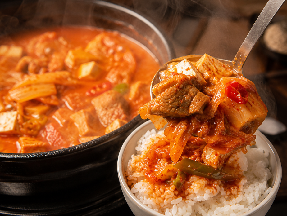 김이 모락모락 나는 진한 국물의 김치찌개를 한 국자 듬뿍 떠서 갓 지은 밥 위에 올리는 모습.