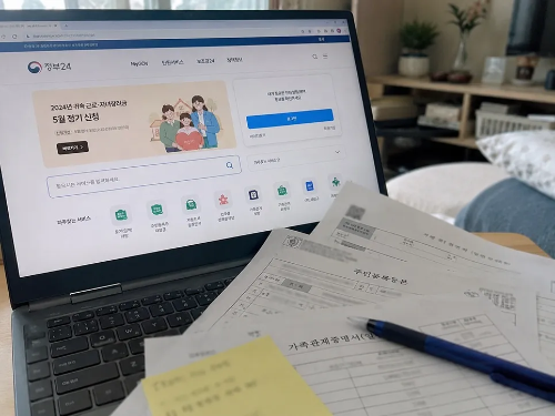 노트북 화면에 정부24 홈페이지가 열려 있고 그 앞 테이블에 인쇄된 주민등록등본과 가족관계증명서 서류, 파란색 볼펜, 노란색 메모지가 놓여 있는 모습