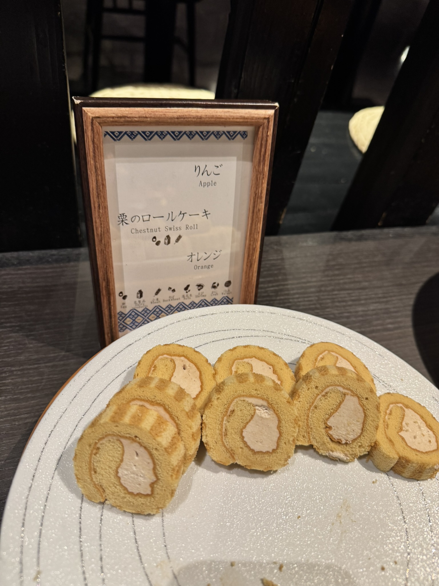 조식 디저트로 제공되는 '밤 롤케이크(Chestnut Swiss Roll)'. 부드러운 시트 속에 밤 크림이 듬뿍 들어간 롤케이크가 접시에 담겨 있고, 뒤쪽 액자에는 오늘의 과일 메뉴인 사과와 오렌지가 안내되어 있음.
