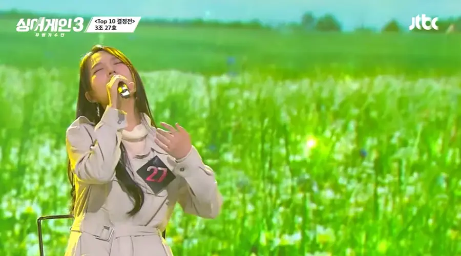 싱어게인3- 초록색 풀밭을 배경으로 흰옷을 입은 27호가 긴머리를 늘어뜨린채 하늘을 바라보며 노래하는 모습