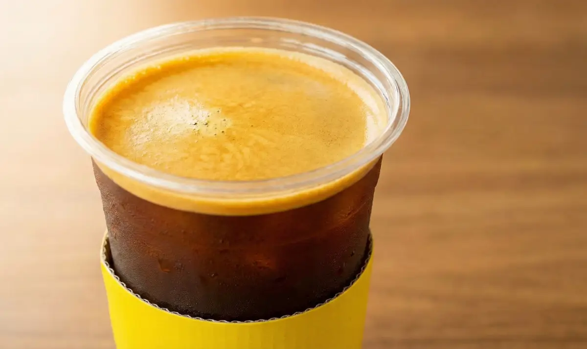 컴포즈커피의 시그니처인 아이스 아메리카노가 노란색 컵홀더에 담겨 있는 모습. 풍부한 크레마와 시원한 얼음이 강조됨.