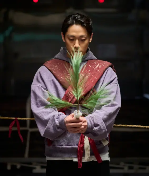 파묘- 어두운 배경 보라색옷에 빨간띠를 몸에 X자로 두루고 나뭇가지를 두손으로 들고있는 이도현