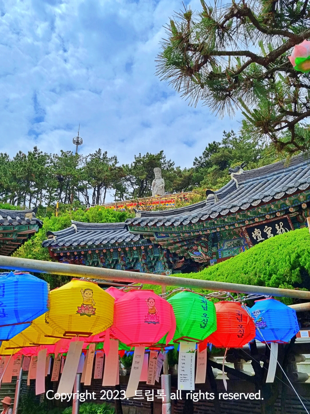 파랑&amp;#44; 노랑&amp;#44; 연두&amp;#44; 빨강 등의 연등이 달려있는 위에 기와지붕이 보이고 그 위에 돌로 된 불상이 있다.