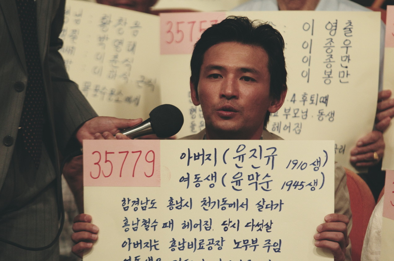 이산가족 찾기 방송 현장에서 젊은 덕수가 가족 이름과 정보가 적힌 피켓을 들고 마이크 앞에 서 있는 장면