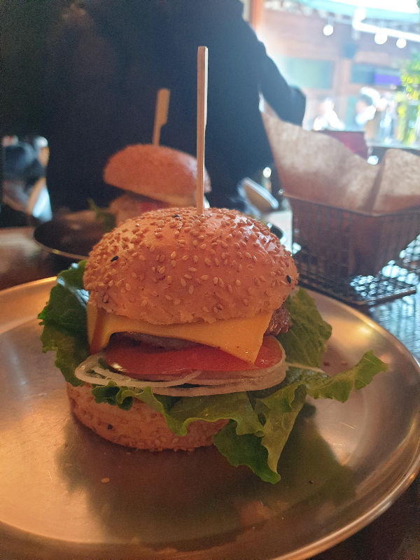 패티, 양파, 상치, 토마토, 치즈가 보이는 햄버거. 번에 깨가 뿌려져 있다.