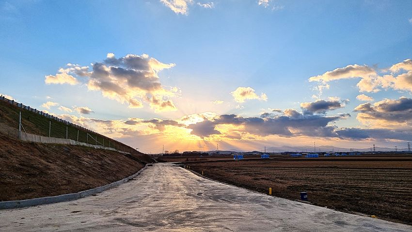 파란 하늘에 강렬한 빛 내림, 역광에 회색 구름들, 왼쪽에 둑방길, 우측에 들판,