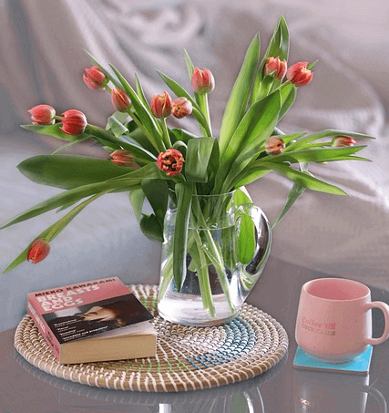 투명한 병에 분홍색 튤립꽃들이 가득 있다. 주변엔 책과 분홍색 머그잔이 있다.