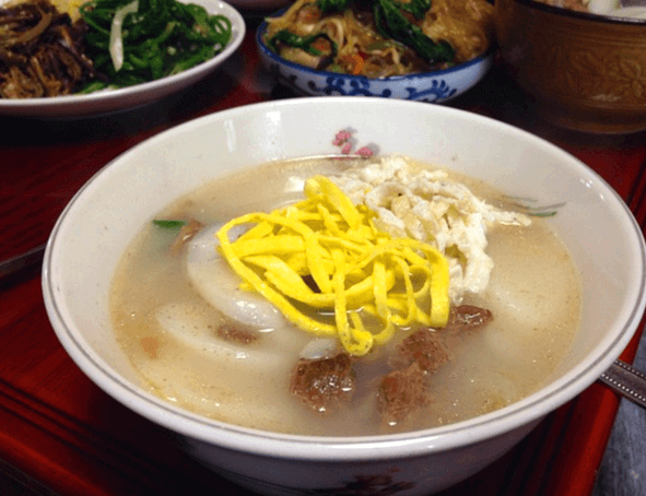 하얀 그릇에 떡국이 담겨있다. 하얀 떡들과 노란 달걀 고명들이 올려져있고 국물에는 소고기가 떠 있다.