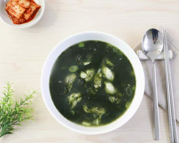 하얀 대접에 담긴 진한 녹색의 매생이국과 굴, 그 옆에 정갈하게 놓인 수저와 김치 반찬이 있는 상차림 이미지