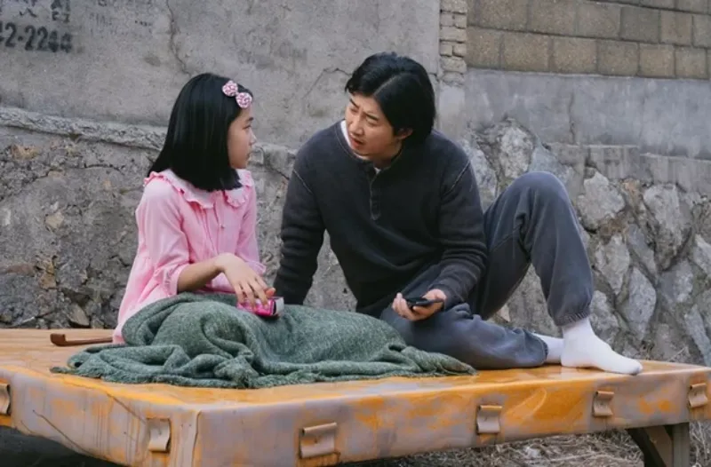 어느 시멘트 담벼락을 배경으로 노란 평상마루에 앉아 있는 드라마 유괴의 날의 배우 윤계상과 유나의 등장 장면