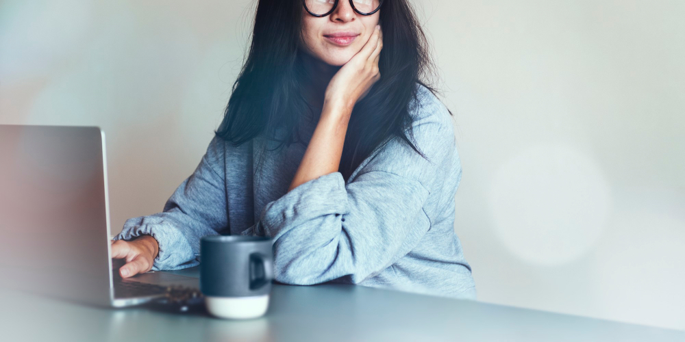 회색 스웨터를 입고 안경을 쓴 여성이 노트북 앞에서 한 손으로 턱을 괴고 생각에 잠긴 채 블로그 포스팅을 작성하고 있으며, 테이블 위에는 커피 컵이 놓여 있다