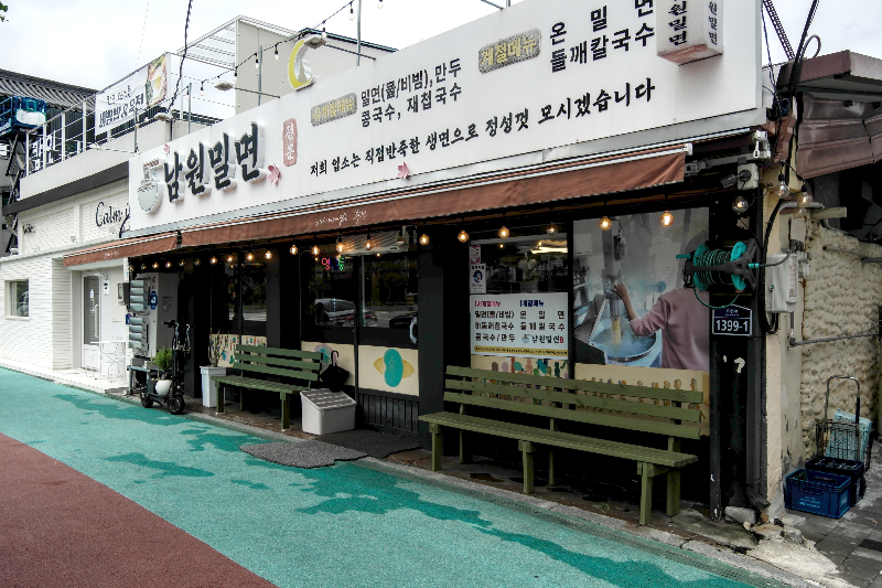 흰색 바탕에 검은 글씨로 남원 밀면이라고 쓰인 간판, 출입문 양 옆 초록색 벤치가 있다.