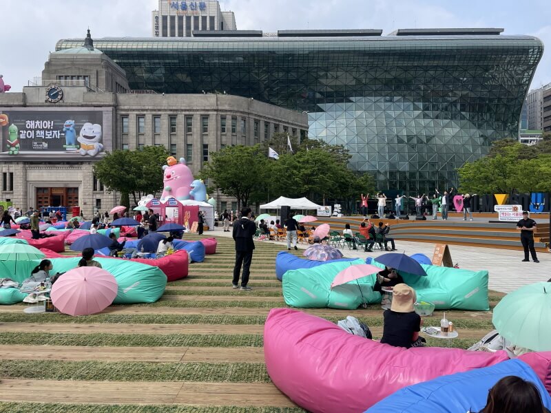 서울도서관과 서울시청 건물을 배경으로 펼쳐진 문화가 흐르는 서울광장 낮 공연 풍경, 시민들이 빈백과 우산을 들고 휴식 중
