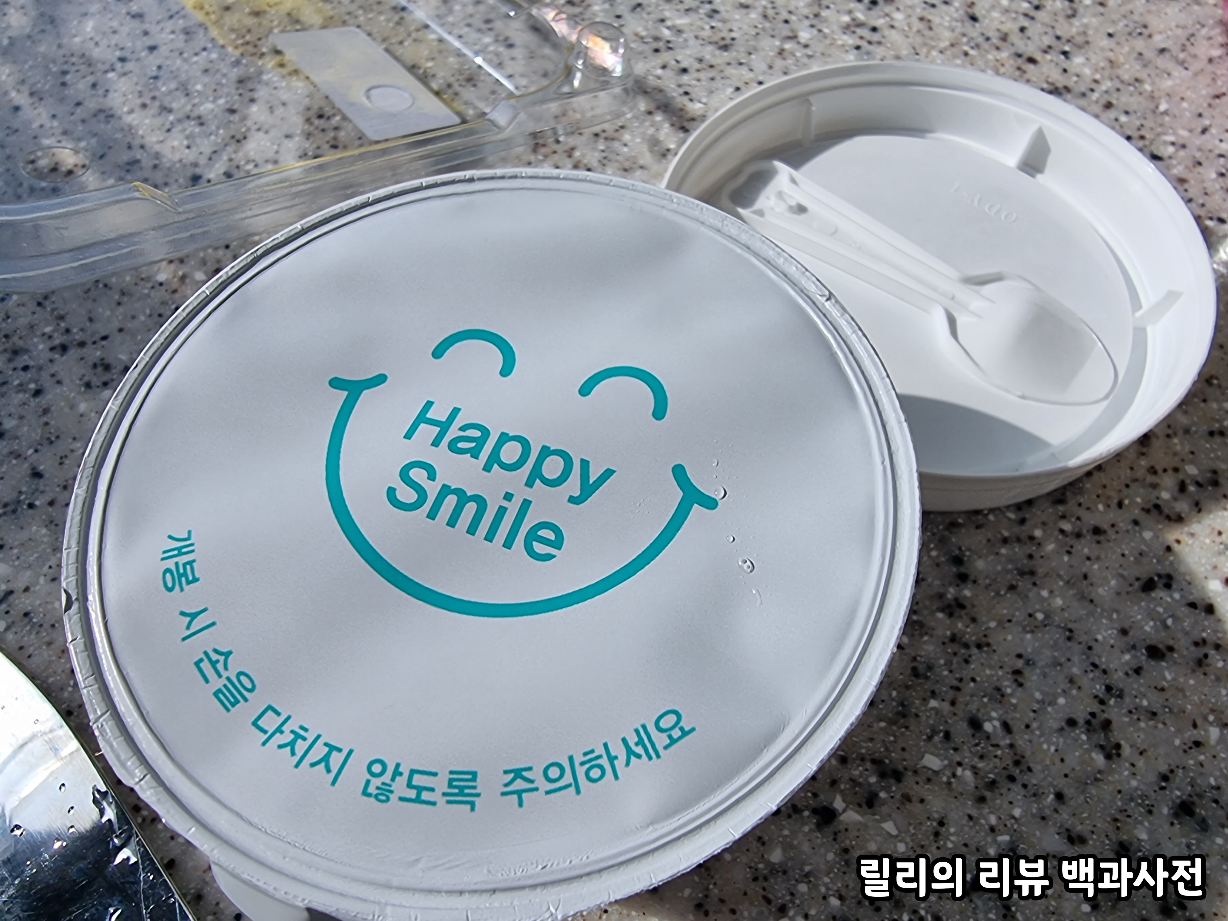 뚜껑을 연 그릭요거트 사진&#44; 뚜껑 안쪽에는 작은 숟가락이 붙어있고 그릭요거트는 Happy Smile&#44; 개봉 시 손을 다치지 않도록 주의하세요 라는 씰이 붙어있다.