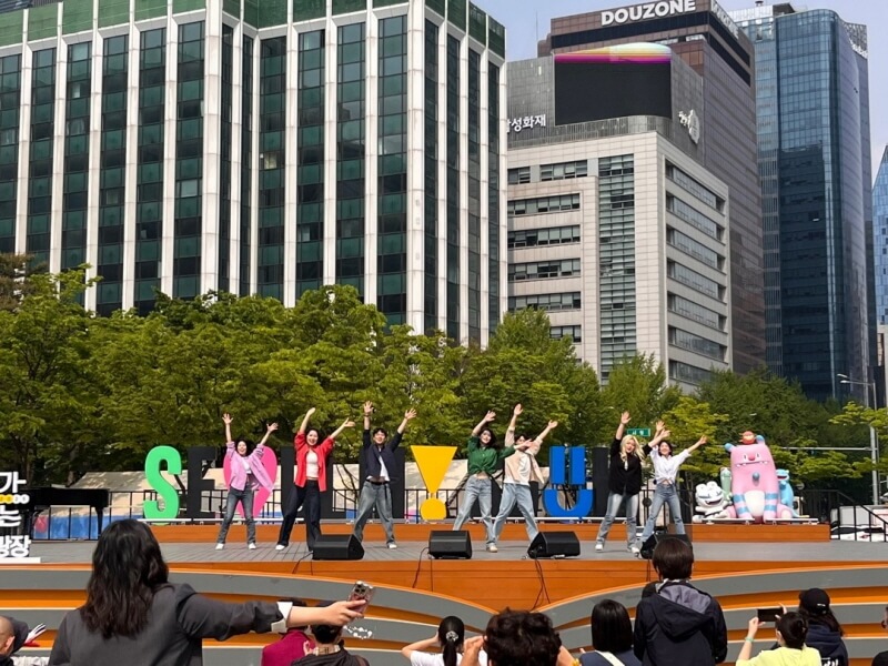 도심 빌딩 숲 속 서울광장 무대에서 단체 댄스를 선보이는 퍼포먼스 팀, 밝은 햇살 아래 관객들이 함께 호응하는 모습